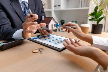 Bir emlakçı ve bir müşteri, emlak satın alma toplantısı sırasında bir konut sözleşmesi belgesini tartışıyor ve ona ev sahipliği hakkında anlaşma detayları ve mali müzakereleri gösteriyorlar.