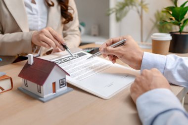 Bir emlakçı ve bir müşteri, emlak satın alma toplantısı sırasında bir konut sözleşmesi belgesini tartışıyor ve ona ev sahipliği hakkında anlaşma detayları ve mali müzakereleri gösteriyorlar.