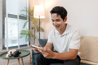 Genç bir adam evde mobil oyun oynarken akıllı telefonunu neşeli bir ifadeyle tutarak koltuğa rahatça oturur. Rahatlamış atmosfer gündelik eğlence ve boş zamanı yansıtıyor..