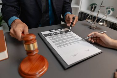 Takım elbiseli bir erkek avukat, yasal bir belgedeki imza bölgesine işaret ederek sözleşmenin detaylarını kadın bir müşteriyle tartışıyor. Ofis masasında hukuk ve adaleti sembolize eden bir tokmak var..
