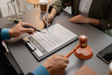Takım elbiseli bir erkek avukat, yasal bir belgedeki imza bölgesine işaret ederek sözleşmenin detaylarını kadın bir müşteriyle tartışıyor. Ofis masasında hukuk ve adaleti sembolize eden bir tokmak var..