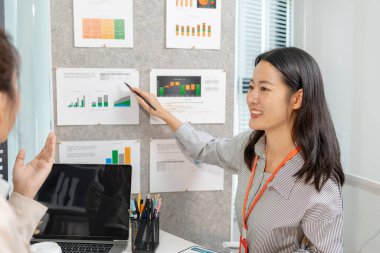 İki Asyalı iş kadını, modern bir ofiste şirket performans raporlarını tartışırken gülümsüyor. Takım çalışması, planlama ve iş birliğini gösteren yazılı belgelerden elde edilen verileri analiz ederler.
