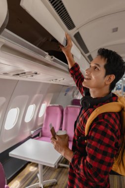Genç adam eşyalarını bir uçağın üst kompartımanına koyarak bir uçuş için hazırlanıyor. Neşeli ve seyahat macerasına hazır görünüyor, elinde bir sırt çantası ve pasaportuyla..