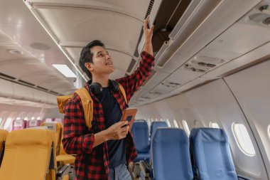 Genç adam eşyalarını bir uçağın üst kompartımanına koyarak bir uçuş için hazırlanıyor. Neşeli ve seyahat macerasına hazır görünüyor, elinde bir sırt çantası ve pasaportuyla..