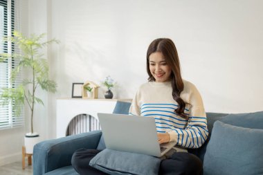 Genç bir kadın kanepede rahat bir şekilde oturuyor, dizüstü bilgisayar kullanıyor, odaklanarak ve sevinçle gülümsüyor. İşiyle ya da internet iletişimiyle meşgul görünüyor, sıcak bir yaşam tarzı konsepti yaratıyor..