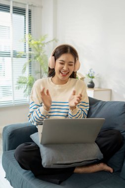 Kanepede oturan genç bir kadın, kulaklık takıyor, dizüstü bilgisayarının önünde alkışlarken mutlu bir şekilde gülümsüyor, evdeki online bir aktivite sırasında neşe ve heyecan gösteriyor..