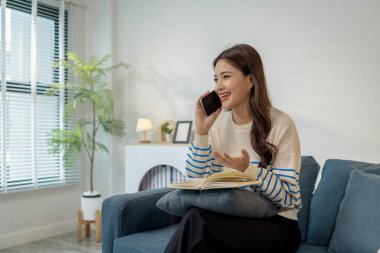 Kanepede rahat bir şekilde oturan genç bir kadın, kucağında bir not defteriyle akıllı bir telefonla gülümserken dijital iletişim ve not alma arasındaki çoklu görevi gösteriyor..