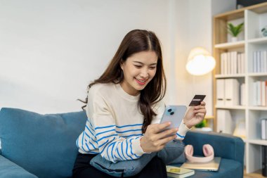 Genç bir kadın elinde akıllı telefon ve kredi kartıyla kanepede oturuyor. Çevrimiçi alışveriş yaparken gülümsüyor. Evde alışveriş yaparken rahat ve kendinden emin görünüyor..