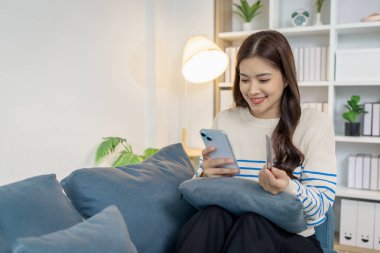 Genç bir kadın elinde akıllı telefon ve kredi kartıyla kanepede oturuyor. Çevrimiçi alışveriş yaparken gülümsüyor. Evde alışveriş yaparken rahat ve kendinden emin görünüyor..