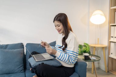Genç bir kadın elinde bir kredi kartı tutarken bir dizüstü bilgisayar kullanarak kanepede rahatça oturuyor ve internet üzerinden ödeme yapıyormuş gibi görünüyor. Memnuniyetle gülümser, rahatlığı ve rahatlığı yansıtır..