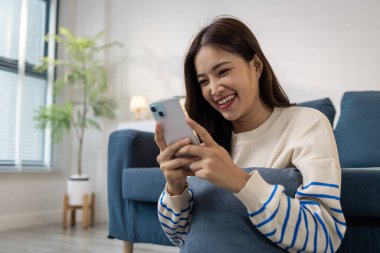 Genç bir kadın yerde minderiyle oturuyor, akıllı telefonunu kullanırken mutlu bir şekilde gülümsüyor. Rahatlamış ve neşeli görünüyor. Evde pozitif enerjisiyle vakit geçiriyor..