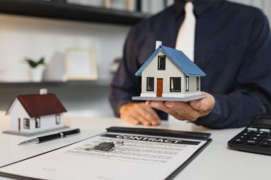 Bir işadamı ev modelleri, anahtarlar ve sözleşme belgeleri, emlak yatırımları ve ipotek anlaşmalarının altını çizerek ev sahipliği, finans ve emlak başarısını sembolize ediyor.