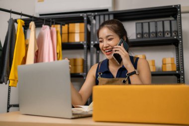 Gülümseyen genç bayan girişimci cep telefonuyla konuşuyor ve ev ofisinden dizüstü bilgisayarla çalışıyor, çevrimiçi giyim mağazasının müşteri siparişlerini yönetiyor ve sevkiyat ve teslimat için ürünler hazırlıyor..