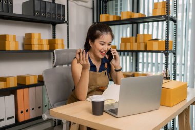 Gülümseyen genç bayan girişimci cep telefonuyla konuşuyor ve ev ofisinden dizüstü bilgisayarla çalışıyor, çevrimiçi giyim mağazasının müşteri siparişlerini yönetiyor ve sevkiyat ve teslimat için ürünler hazırlıyor..