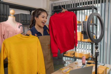 Genç bayan girişimci, ev ofisinden kıyafet satmak için yaşarken gülümsüyor. Moda ürünleri sunar, izleyicilerle iletişim kurar ve canlı seans sırasında online mağazasını yönetir..