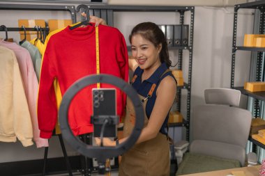 Genç bayan girişimci, ev ofisinden kıyafet satmak için yaşarken gülümsüyor. Moda ürünleri sunar, izleyicilerle iletişim kurar ve canlı seans sırasında online mağazasını yönetir..