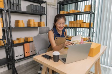 Evde dizüstü bilgisayarı üzerinde çalışırken gülümseyen genç bir girişimci. Etrafı kıyafet ve paketlerle çevrili. Bir internet mağazası işletiyor, müşterilerle iletişim kuruyor ve e-ticaret satışlarına hazırlanıyor..