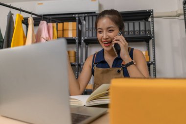 Genç bayan girişimci telefonda konuşurken gülümsüyor ve evde dizüstü bilgisayar kullanıyor. Bir internet mağazası işletiyor, siparişleri kontrol ediyor ve e-ticaret işi için müşterilerle iletişim kuruyor..
