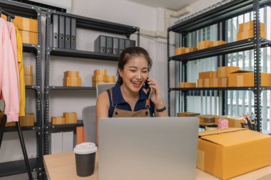 Genç bayan girişimci telefonda konuşurken gülümsüyor ve evde dizüstü bilgisayar kullanıyor. Bir internet mağazası işletiyor, siparişleri kontrol ediyor ve e-ticaret işi için müşterilerle iletişim kuruyor..