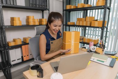 Genç bayan girişimci elinde bir paket tutarken gülümsüyor ve evinde dizüstü bilgisayar kullanıyor. Bir internet mağazası işletiyor, teslimatları hazırlıyor ve müşterilerle e-ticaret için iletişim kuruyor..