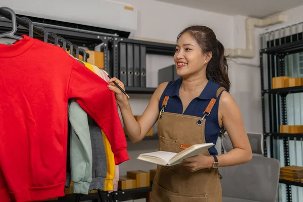 Neşeli genç bayan girişimci elinde defter tutarken envanterini kontrol edip teftiş ediyor, çevrimiçi moda mağazasını yönetiyor, iş yerinde ürün ve siparişleri hazırlıyor..