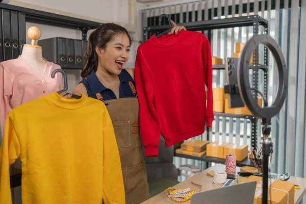 Genç bayan girişimci, ev ofisinden kıyafet satmak için yaşarken gülümsüyor. Moda ürünleri sunar, izleyicilerle iletişim kurar ve canlı seans sırasında online mağazasını yönetir..