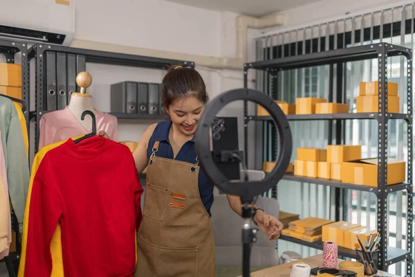 Genç bayan girişimci, ev ofisinden kıyafet satmak için yaşarken gülümsüyor. Moda ürünleri sunar, izleyicilerle iletişim kurar ve canlı seans sırasında online mağazasını yönetir..