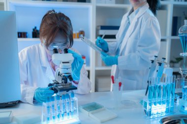 Beyaz laboratuvar önlüklü iki kadın bilim adamı, takım çalışmasını ve modern bilimsel araştırma ortamını temsil eden mikroskoplar, cam eşyalar ve mavi test tüpleriyle parlak bir laboratuvarda birlikte çalışıyorlar..
