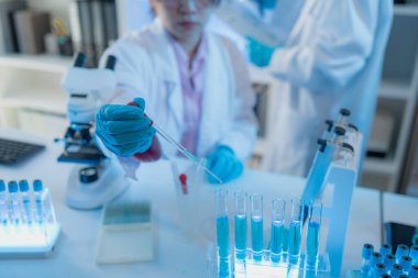 Beyaz laboratuvar önlüklü iki kadın bilim adamı, takım çalışmasını ve modern bilimsel araştırma ortamını temsil eden mikroskoplar, cam eşyalar ve mavi test tüpleriyle parlak bir laboratuvarda birlikte çalışıyorlar..