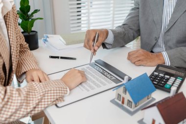 Bir emlakçı ve müşteri emlak satın alma seçeneklerini tartışıyor, sözleşmeyi gözden geçiriyor, kredi şartlarını hesaplıyor ve ev modellerini konut yatırımı kararının bir parçası olarak değerlendiriyor..