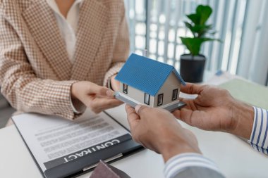 Bir emlakçı, şartları açıklamak için ev modelleri ve hesap makinesi kullanarak bir müşteriyle emlak sözleşmelerini tartışır. Sahne, müzakere, karar verme ve mali planlamayı vurguluyor.