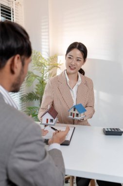 Bir emlakçı, şartları açıklamak için ev modelleri ve hesap makinesi kullanarak bir müşteriyle emlak sözleşmelerini tartışır. Sahne, müzakere, karar verme ve mali planlamayı vurguluyor.