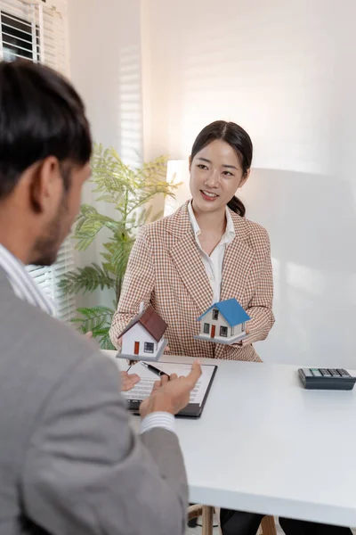 Bir emlakçı, şartları açıklamak için ev modelleri ve hesap makinesi kullanarak bir müşteriyle emlak sözleşmelerini tartışır. Sahne, müzakere, karar verme ve mali planlamayı vurguluyor.