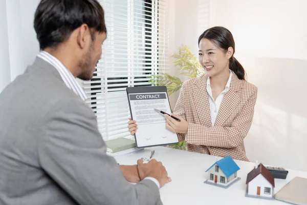 Bir emlakçı, şartları açıklamak için ev modelleri ve hesap makinesi kullanarak bir müşteriyle emlak sözleşmelerini tartışır. Sahne, müzakere, karar verme ve mali planlamayı vurguluyor.