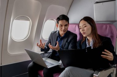 Asyalı iş adamları uçakta uçarken iş konuşuyorlar. Kadın elinde bir klasör ve kalem tutarken adam bir şey açıklarken el kol hareketi yapıyor..