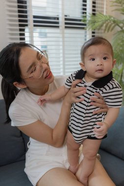 Genç bir anne, bebeğini sevgiyle kucaklar ve bir kanepede emzirir. Erken çocukluk ve aile hayatının anne bakımını, bağlarını ve besleyici anlarını sembolize eder..