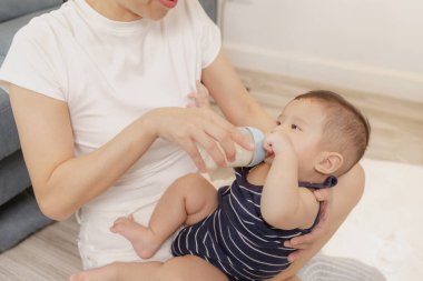 Bir anne çocuğunu kucağında tutarken sevgiyle bebeğini bir şişe sütle besler. Sahne ebeveynlik, bakım ve annelik ilişkisinin sıcaklığını vurguluyor.