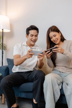 Neşeli bir Asyalı çift kanepede birlikte oturuyor, gezici oyunlar oynuyor ve rahat bir oturma odasında mutlu bir şekilde gülüyor. Boş zamanlarını ve keyifli dostça rekabetlerini severler..