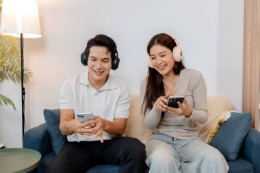 Kablosuz kulaklık takan neşeli bir adam ve kadın, rahat bir kanepede oturup kahkaha atarken ve evde rahat bir atmosferi paylaşırken akıllı telefonlarıyla müzik dinlemenin keyfini çıkarırlar..