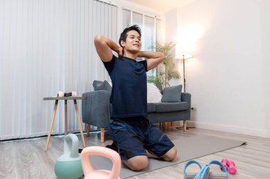 Evinde, rahat bir oturma odasında egzersiz yapan, sağlığına ve sağlığına odaklanan genç bir adam. Dengeli bir yaşam tarzını destekleyen basit spor aletleriyle çevrili günlük egzersiz rutinini seviyor..