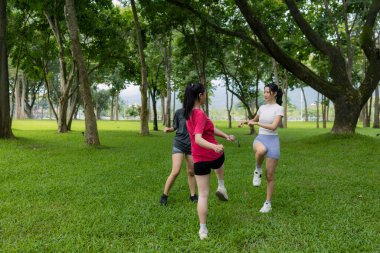 Üç kadın yemyeşil bir parkta birlikte egzersiz yapıyor, uzun ağaçların arasında aktif bir açık hava egzersizinin tadını çıkarıyorlar. Neşeli ve odaklanmış görünüyorlar. Arkadaşlığı, formda kalmayı ve sağlıklı bir yaşam tarzını teşvik ediyorlar..