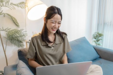 Genç bir kadın elinde dizüstü bilgisayarla kanepede oturuyor. Ekrana bakıp gülümsüyor. Rahatlamış ve meşgul görünüyor, çevrimiçi iletişimden, uzaktan çalışmadan ya da evde dijital öğrenmeden hoşlanıyor..