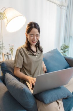 Genç bir kadın elinde dizüstü bilgisayarla kanepede oturuyor. Ekrana bakıp gülümsüyor. Rahatlamış ve meşgul görünüyor, çevrimiçi iletişimden, uzaktan çalışmadan ya da evde dijital öğrenmeden hoşlanıyor..