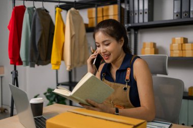 Not defteri tutarken cep telefonuyla konuşan mutlu genç bayan girişimci, çevrimiçi giyim mağazası işini yönetiyor, müşteri siparişlerini koordine ediyor ve merkez ofisten sevkiyatları hazırlıyor..