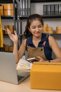 Akıllı telefon ve kalem tutan düşünceli genç bayan girişimci, müşteri siparişlerini dikkatle gözden geçiriyor ve ev ofisinden onun çevrimiçi giyim mağazası işletmelerini yönetiyor..