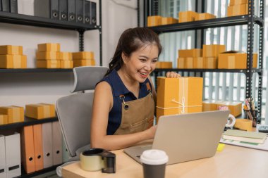 Genç bayan girişimci elinde bir paket tutarken gülümsüyor ve evinde dizüstü bilgisayar kullanıyor. Bir internet mağazası işletiyor, teslimatları hazırlıyor ve müşterilerle e-ticaret için iletişim kuruyor..