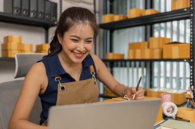 Genç bayan girişimci bir paket etiketinin üzerine bir nakliye adresi yazarak ev ofisine teslimat için bir paket hazırlıyor. Online bir iş yürütüyor ve müşterilere sevkiyat düzenliyor..