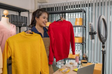 Genç bayan girişimci, ev ofisinden kıyafet satmak için yaşarken gülümsüyor. Moda ürünleri sunar, izleyicilerle iletişim kurar ve canlı seans sırasında online mağazasını yönetir..