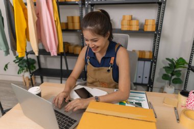 Evde dizüstü bilgisayarı üzerinde çalışırken gülümseyen genç bir girişimci. Etrafı kıyafet ve paketlerle çevrili. Bir internet mağazası işletiyor, müşterilerle iletişim kuruyor ve e-ticaret satışlarına hazırlanıyor..