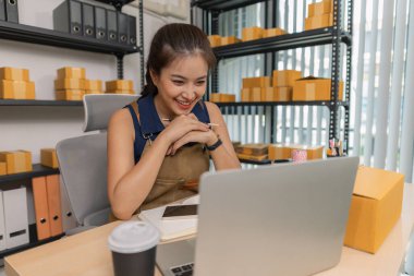 Evde dizüstü bilgisayarı üzerinde çalışırken gülümseyen genç bir girişimci. Etrafı kıyafet ve paketlerle çevrili. Bir internet mağazası işletiyor, müşterilerle iletişim kuruyor ve e-ticaret satışlarına hazırlanıyor..